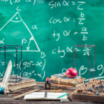 Mathematical implements on a wooden table with scribbled equations on a greenboard.