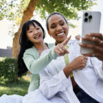 Two young women, one African American and one Asian, happily pose for a selfie while enjoying a picnic in a park.