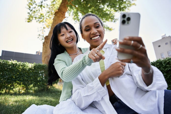 Two young women, one African American and one Asian, happily pose for a selfie while enjoying a picnic in a park.