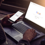 A person using a smartphone and laptop with Google homepage on display.