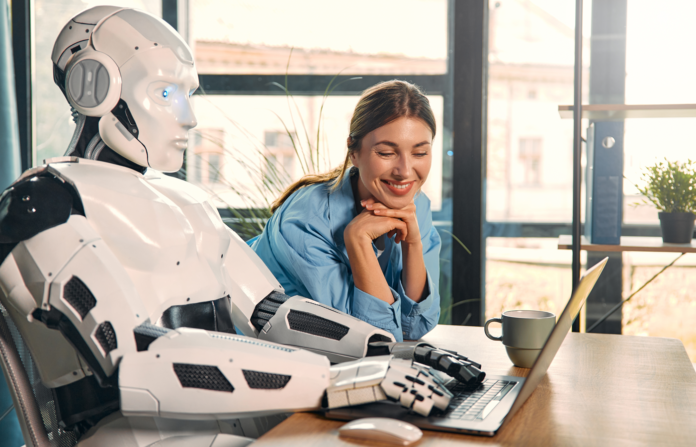 Robot and woman working on laptop in office.