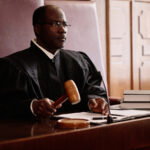 Experienced male judge with wooden gavel over sounding block sitting by desk.