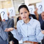 Shot of a group of businesspeople holding up signs with question marks on them during a work presentation while their colleague looks confused.