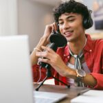 Stock photo of a person using a microphone perhaps to record a podcast and looking at a laptop.