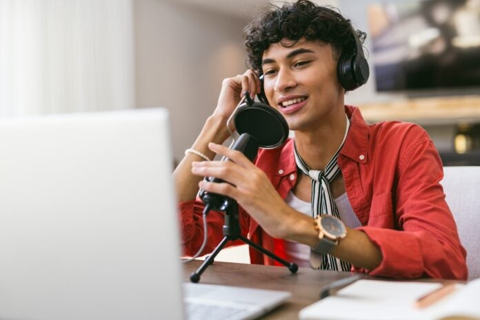 Stock photo of a person using a microphone perhaps to record a podcast and looking at a laptop.
