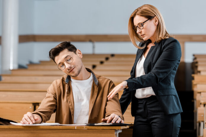 Female university teacher looking at male student in glasses writing exam in classroom.