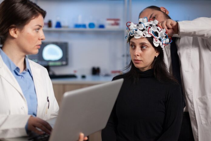 Stock photo of what appears to be a neurologist showing tomography to a patient while a researcher is adjusting an EEG headset.