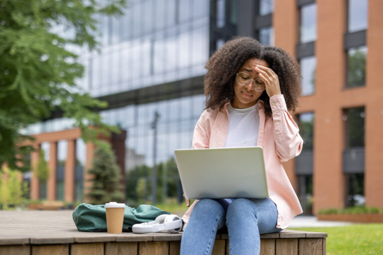 Stressed african american woman student working on laptop on campus. She appears frustrated and exhausted, sitting outside university building with coffee and backpack, showing fatigue with studies.